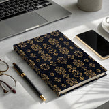 Dark blue hard cover journal with gold baroque pattern, pen, glasses, phone, and coffee.