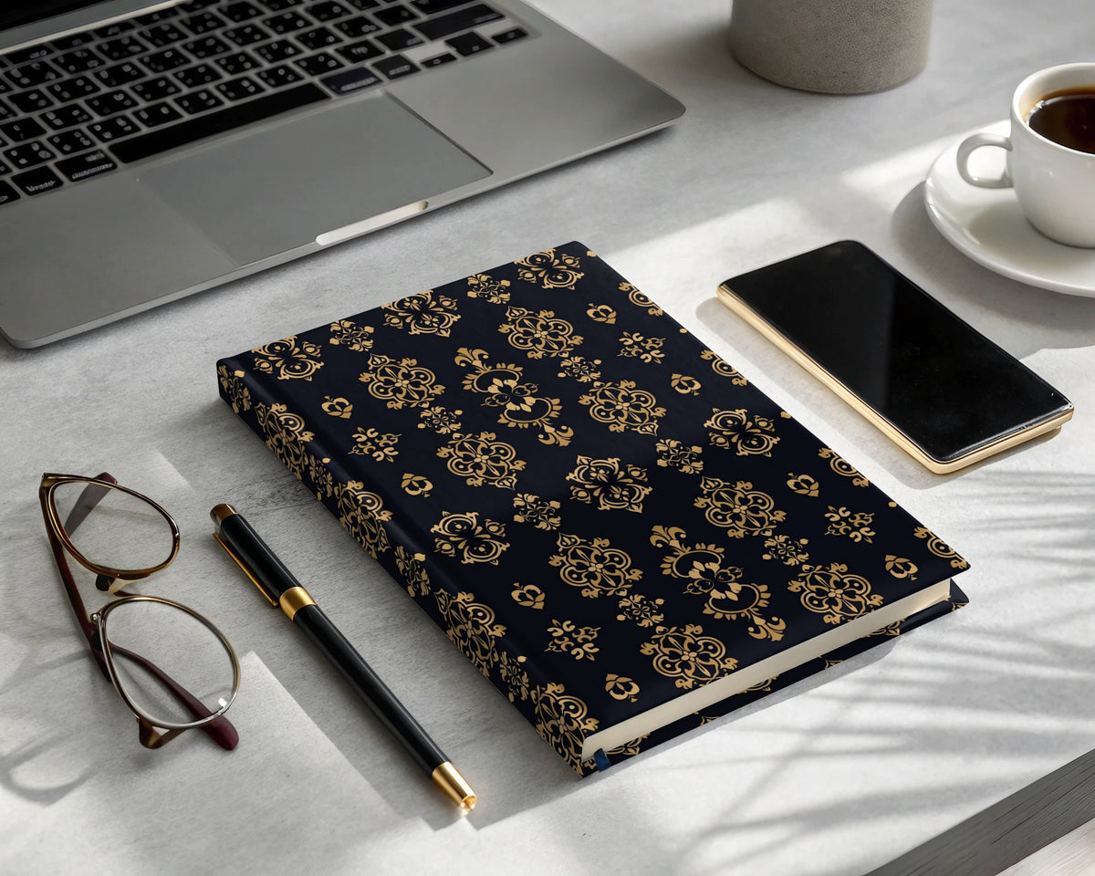 Dark blue hard cover journal with gold baroque pattern, pen, glasses, phone, and coffee.