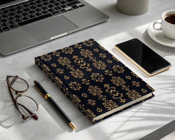 Dark blue hard cover journal with gold baroque pattern, pen, glasses, phone, and coffee.