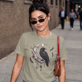 A woman wearing an olive green graphic t-shirt with a black raven and crescent moon design, styled with a brown leather shoulder bag and sunglasses, standing on a city sidewalk.