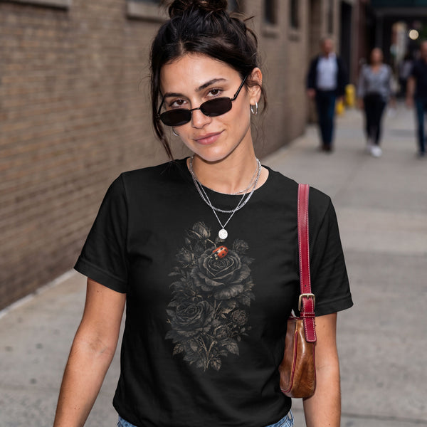 A woman wearing a black graphic t-shirt with a gothic key and rose design, styled with sunglasses, layered necklaces, and a brown leather shoulder bag, standing on a city sidewalk.