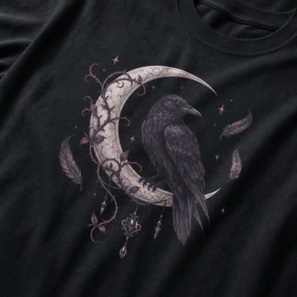 A close-up of a black t-shirt featuring a detailed graphic print of a raven perched on a crescent moon, decorated with dark purple vines, feathers, and small stars.