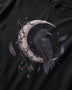A close-up of a black t-shirt featuring a detailed graphic print of a raven perched on a crescent moon, decorated with dark purple vines, feathers, and small stars.