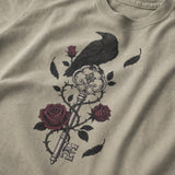 A close-up of a beige t-shirt featuring a graphic print of a black raven perched on an ornate key with dark red roses and black leaves.