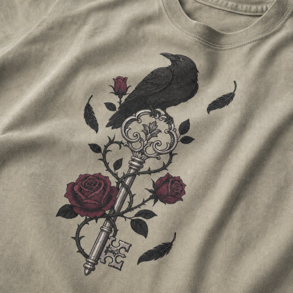 A close-up of a beige t-shirt featuring a graphic print of a black raven perched on an ornate key with dark red roses and black leaves.