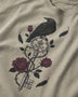 A close-up of a beige t-shirt featuring a graphic print of a black raven perched on an ornate key with dark red roses and black leaves.