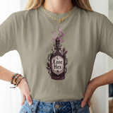 A woman wearing a sage green t-shirt with a gothic-style 'Love Hex' potion bottle graphic, paired with blue jeans and gold jewelry.