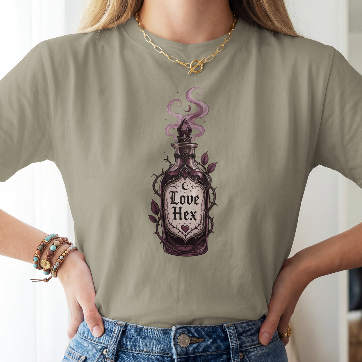 A woman wearing a sage green t-shirt with a gothic-style 'Love Hex' potion bottle graphic, paired with blue jeans and gold jewelry.