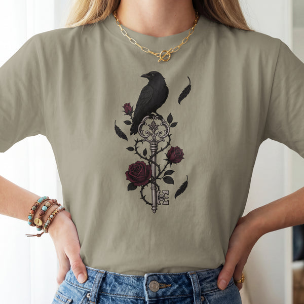 A woman wearing a sage green t-shirt with a gothic-style graphic of a black raven perched on a vintage key with dark red roses. The model is also wearing blue jeans, a gold chain necklace, and multiple beaded bracelets.