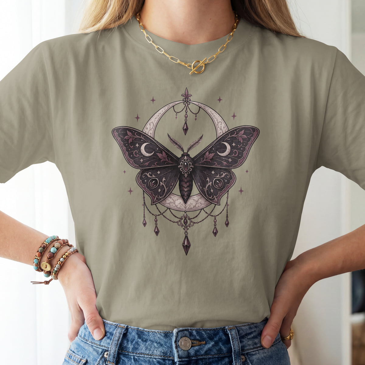 A woman wearing a sage green t-shirt with a detailed graphic of a dark purple moth inside a crescent moon and dreamcatcher design, paired with blue jeans.