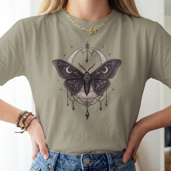 A woman wearing a sage green t-shirt with a detailed graphic of a dark purple moth inside a crescent moon and dreamcatcher design, paired with blue jeans.