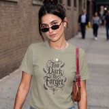 A woman wearing a sage green graphic t-shirt with the text 'Too Dark To Forget' in a gothic font, styled with black sunglasses, layered necklaces, and a brown leather shoulder bag, set against a blurred city street background.