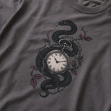 A close-up of a dark gray t-shirt featuring a graphic print of a black snake wrapped around a vintage pocket watch, accented with barbed wire and muted red-purple leaves.