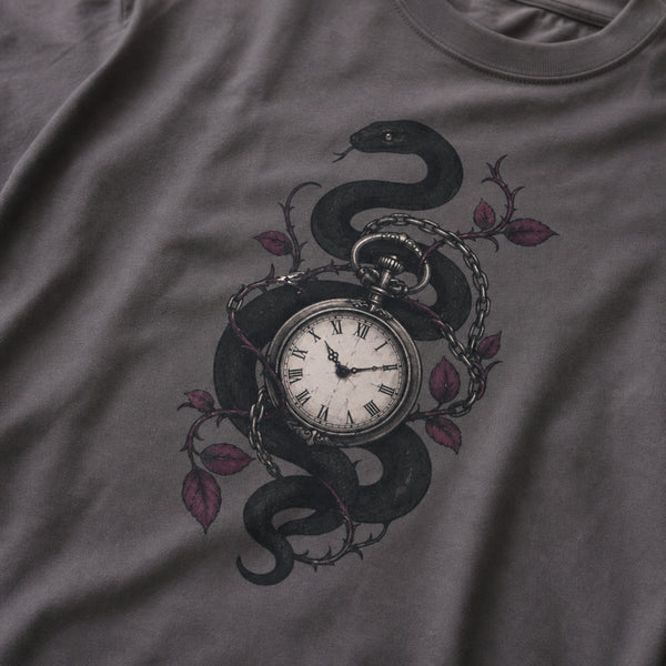 A close-up of a dark gray t-shirt featuring a graphic print of a black snake wrapped around a vintage pocket watch, accented with barbed wire and muted red-purple leaves.