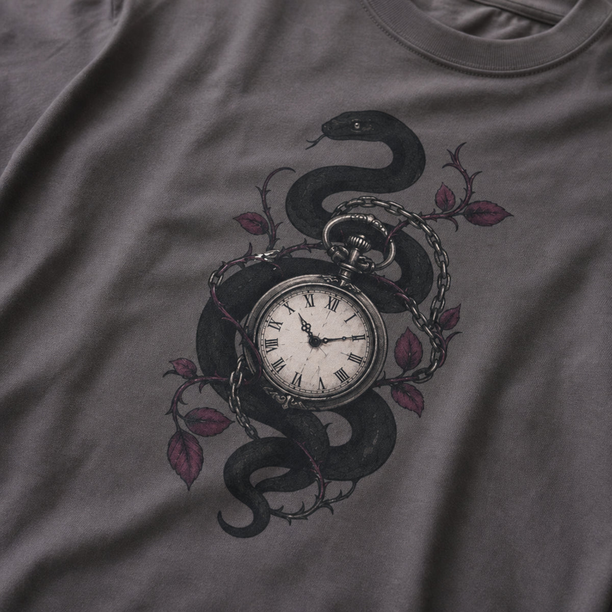 A close-up of a dark gray t-shirt featuring a graphic print of a black snake wrapped around a vintage pocket watch, accented with barbed wire and muted red-purple leaves.