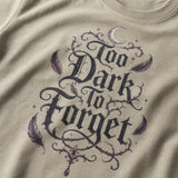 Close-up of a beige t-shirt featuring a gothic-style graphic print that reads 'Too Dark to Forget' in black lettering, decorated with purple feathers, thorny vines, and a small crescent moon.