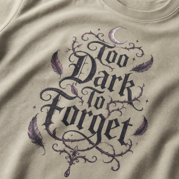 Close-up of a beige t-shirt featuring a gothic-style graphic print that reads 'Too Dark to Forget' in black lettering, decorated with purple feathers, thorny vines, and a small crescent moon.