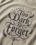 Close-up of a beige t-shirt featuring a gothic-style graphic print that reads 'Too Dark to Forget' in black lettering, decorated with purple feathers, thorny vines, and a small crescent moon.