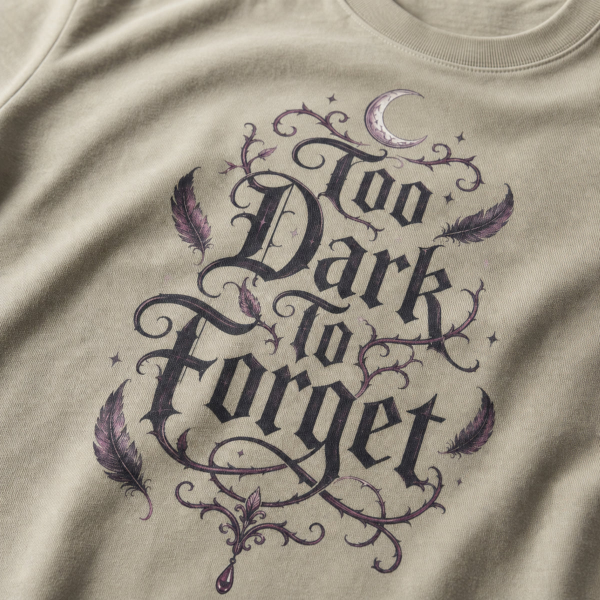 Close-up of a beige t-shirt featuring a gothic-style graphic print that reads 'Too Dark to Forget' in black lettering, decorated with purple feathers, thorny vines, and a small crescent moon.