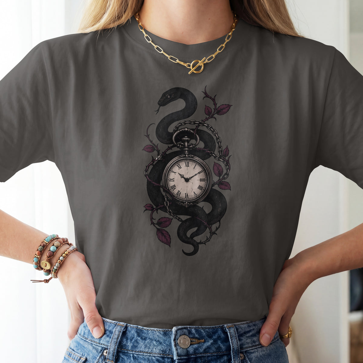 A woman wearing a dark gray t-shirt with a graphic of a black snake wrapped around a pocket watch and purple leaves, paired with blue jeans.