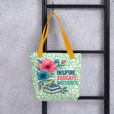 Tote bag with floral design and text 'Inspire. Educate. Motivate.' hanging on a black metal rack against a concrete wall.