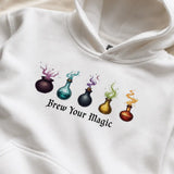 White hoodie with colorful potion bottles and 'Brew Your Magic' text on a beige background XS