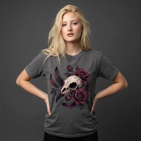A blonde woman modeling a dark gray t-shirt with a graphic print of a bird skull and dark purple roses, posed against a dark gray studio background.