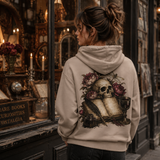 Person wearing a hoodie with a skull and floral design in front of an antique shop window.