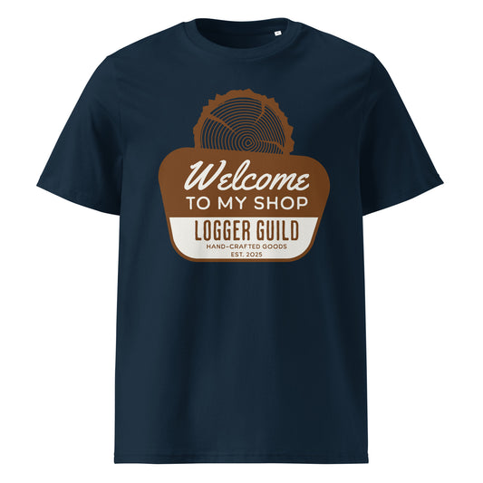 A french navy unisex organic cotton t-shirt featuring a brown and white graphic. The graphic shows a stylized wood slice or saw blade at the top, above a brown banner with 'Welcome TO MY SHOP' in white cursive. Below that, 'LOGGER GUILD' is prominently displayed in white block letters on a white stripe, followed by 'HAND-CRAFTED GOODS' and 'EST. 2025' in smaller brown text. The shirt has a ribbed neck."